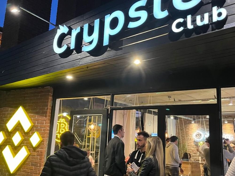 Crypster Club de noche, con el cartel iluminado, el logo de Binance en la pared y gente entrando al salón