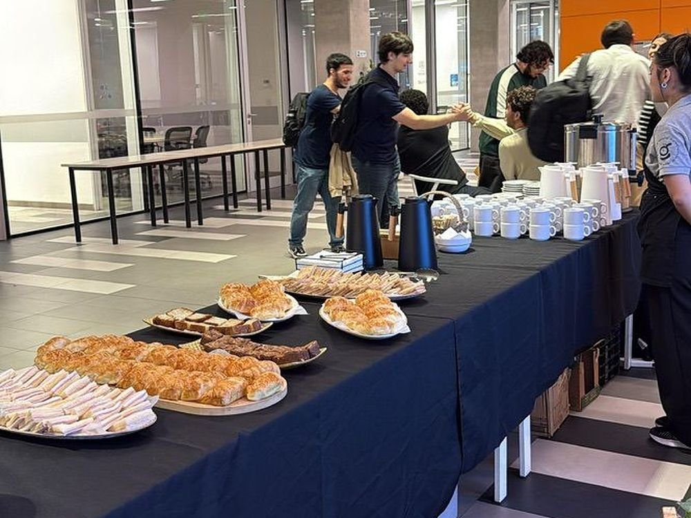 Desayuno de trabajo montado sobre mesa larga, con bandejas de sándwiches, facturas, tazas y termos de café