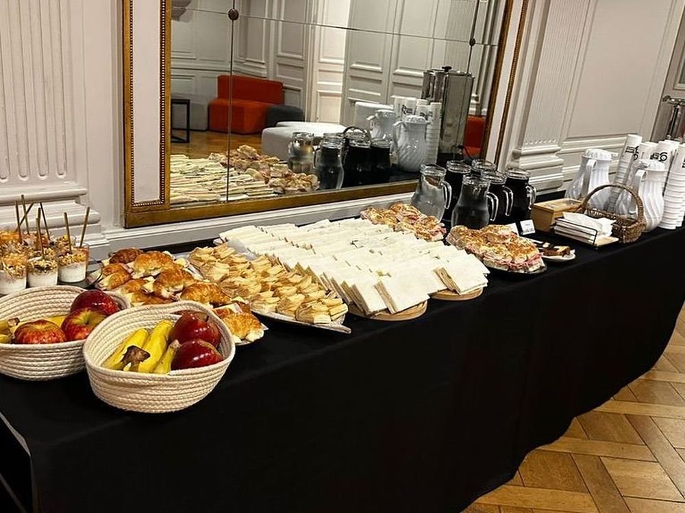 Mesa de coffee break montada con bandejas de sándwiches, facturas, fruta y termos de café